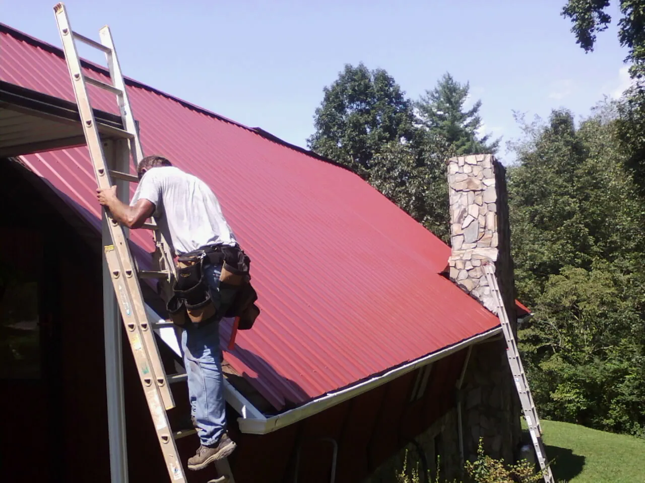 Roofer on a ladder beside a red metal roof and stone chimney — Soffit & Fascia Repair by Roofing Brothers in Cordele